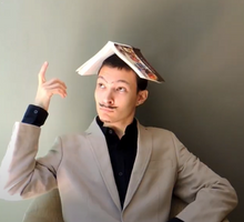 Foto de um homem branco com um livro aberto apoiado na cabeça. Ele  veste roupas sociais, usa um bigode falso, olha para o lado e levanta um dedo. Ele está sentado em frente a uma parede cinza.