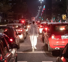 Foto tirada durante a noite na Avenida Paulista, entre duas faixas de carros. Há diversos veículos nas duas faixas, eles estão com as lanternas traseiras acesas, indicando que estão em marcha à ré. Entre eles, há uma pessoa coberta por uma roupa de proteção branca, ela usa máscara. 