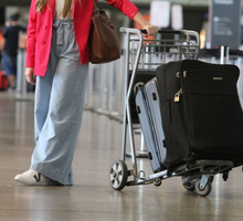 Foto de uma mulher apoiada em um carrinho de bagagens. Ela é branca, tem cabelos longos e veste uma camiseta branca, um blazer rosa e uma calça jeans azul claro. Suas duas malas são escuras. A foto está cortada no meio de seu tronco.