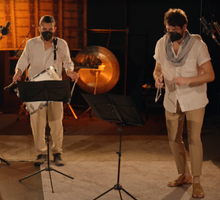 Foto de dois homens tocando instrumentos em um estúdio. O da esquerda faz a percussão e o da direita toca um triângulo. Ambos são brancos, têm cabelos curtos escuros, usam máscara e vestem uma camisa branca e uma calça bege. Eles olham para suas partituras.