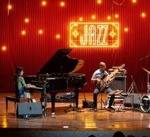  Foto de um concerto de jazz. Há em cima de um palco e madeira uma pianista e um contrabaixista. Ambos estão tocando. Ao fundo, há uma parede vermelha, com pontos luminosos amarelos e um letreiro neon, também amarelo, escrito “jazz”. Parte de uma bateria também faz parte da foto.