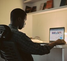 Foto vista por trás de homem usando um laptop. Ele tem a pele preta, cabelos curtos, usa camisa xadrez e sua mão está apoiada perto do computador, que está em cima de uma mesa. Ao fundo, há uma prateleira com livros.