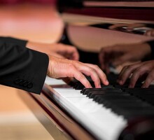 Foto de uma pessoa tocando um piano. Apenas as mãos do musicista aparecem sobre as teclas, que são pretas e brancas. O piano é feito de madeira escura.