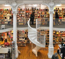 Foto de uma livraria com dois andares. Há algumas pessoas observando os livros nas estantes. As colunas que sustentam o teto e a escada caracol para chegar ao segundo andar são brancas. O chão é feito de tábuas de madeira.