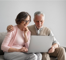  Foto de um casal de idosos. O homem e a mulher estão sentados em um sofá e utilizando juntos um laptop cinza. Ela veste uma blusa rosa e uma calça cinza, enquanto ele usa roupas inteiramente beges. Ele a abraça, e a mão dele está em cima do ombro dela. Os dois sorriem.
