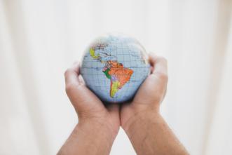 Foto colorida, duas mãos de pessoa branca seguram globo terrestre pequeno, com Continente Americano em evidência