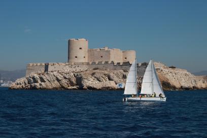 Foto colorida em ambiente externo, com um barco velejando em frente a um forte, no sul da França.