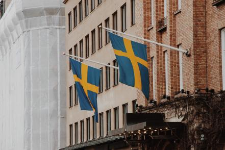 Foto colorida em ambiente externo, de um prédio com duas bandeiras da Suécia penduradas em uma das paredes