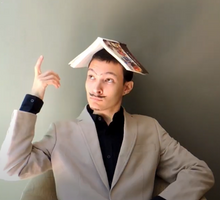 Foto de um homem branco com um livro aberto apoiado na cabeça. Ele veste roupas sociais, usa um bigode falso, olha para o lado e levanta um dedo. Ele está sentado em frente a uma parede cinza.