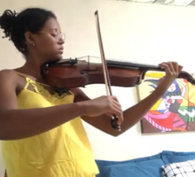 Foto de uma jovem tocando violino. Ela é negra, usa óculos, veste uma blusa amarela e tem seus cabelos escuros presos em um coque. Ao fundo, há uma parede branca, um quadro colorido e travesseiros apoiados na parede.