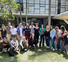 Foto de um grupo de pessoas, que posam sobre um gramado. São homens e mulheres que, em sua maioria, olha para a câmera e sorriem. Atrás deles, um prédio. À esquerda, atrás do grupo e em frente ao prédio, há uma árvore.