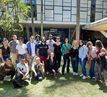 Foto de um grupo de pessoas, que posam sobre um gramado. São homens e mulheres que, em sua maioria, olha para a câmera e sorriem.  Atrás deles, um prédio. À esquerda, atrás do grupo e em frente ao prédio, há uma árvore. 