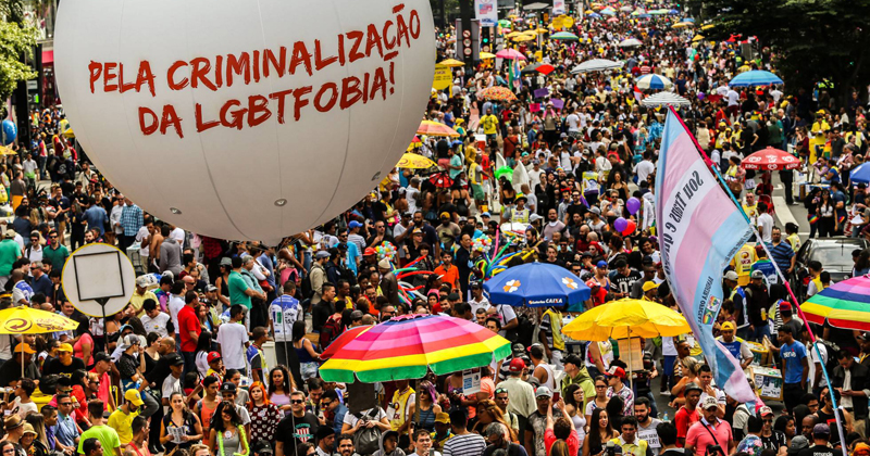 Aglomerado de pessoas na rua em uma manifestação contra a LGBTFobia. Em destaque, no canto esquerdo, um balão de ar branco, com o seguinte texto em vermelho: Pela criminalização da LGBTfobia.