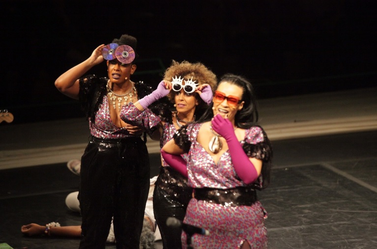 Fotografia colorida de três atrizes negras olhando para algo fora do palco. As três utilizam roupas em cores rosa e preto metálicos e óculos personalizados com CDs. Atrás delas, caída sobre o palco, está uma quarta atriz, de cabelos grisalhos e vestida com roupas brancas.
