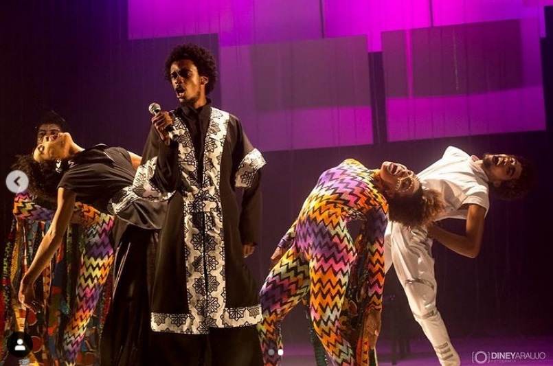 Fotografia colorida mostra um ator negro no centro do palco, cantando e usando roupa preta com detalhes em tecido branco estampado, que lembra tecidos típicos africanos. À sua direita e esquerda atores vestidos com macacões multicoloridos, brancos e pretos dançam.