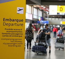 Foto tirada em um aeroporto. Em primeiro plano, à esquerda, está uma placa amarela com o desenho de um avião decolando. Nela, há o título “Embarque” e algumas instruções para os passageiros. Abaixo, o mesmo texto está traduzido para o inglês. Ao fundo, desfocada, há uma movimentação de passageiros com carrinhos de bagagem.