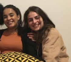Crisley, uma moça negra de pele clara e cabelos pretos presos em rabo de cavalo, usa uma blusa laranja e um casaco preto. Ao lado dela está Ana Gabriela, branca, de cabelos castanhos e lisos e vestida com uma jaqueta nude. Ambas estão sentadas, com os rostos bem próximos uma da outra e sorrindo para a câmera