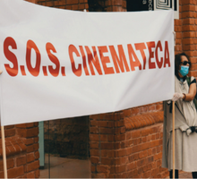 Foto de uma mulher segurando um cartaz de protesto. Ele é branco e tem o texto “S.O.S. CINEMATECA” em vermelho. Ela é branca, veste roupas claras, usa óculos escuros e máscara. Ao fundo, estão os tijolos que formam a fachada do prédio da Cinemateca.