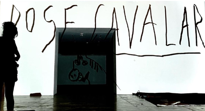 Foto de um espaço em que está escrito no fundo branco em letras pretas “dose cavalar”. No piso cinza, à direita, há uma espécie de lona e, no centro, uma caixa escura com o desenho da cara de um cavalo em traços simples. À esquerda, uma silhueta, aparentemente, de uma mulher.