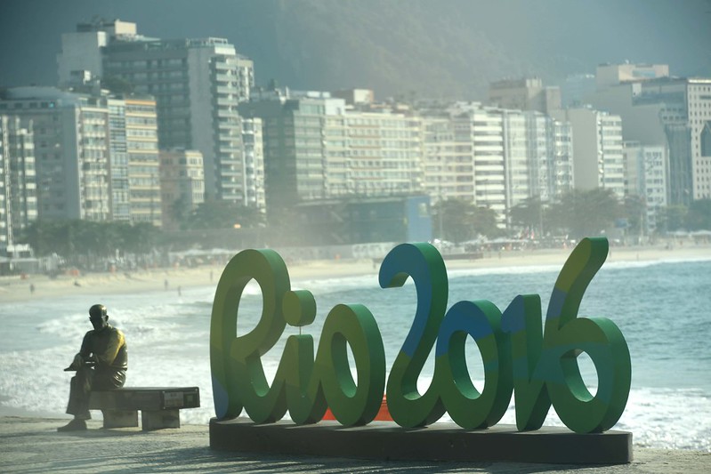 Imagem de monumento em cor verde das palavras Rio 2016, criado para as Olimpíadas do Rio de Janeiro. Ao lado esquerdo, uma estátua em bronze do poeta Carlos Drummond de Andrade sentado em um banco. Ao fundo, a praia de Copacabana e prédios.