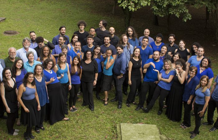 Foto de cerca de 50 pessoas de variadas idades e gêneros, sendo majoritariamente brancas. Elas estão em pé, sorrindo e olhando para cima, de onde é captada a fotografia.  Estão dispostas em três fileiras em semicírculo. A maior parte  utiliza blusas  azuis em diferentes tons  e calças pretas.