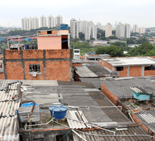 Foto tirada do alto apresentando casas populares em primeiro plano, com muitas paredes sem reboco. Ao fundo, algumas árvores e prédios distantes.