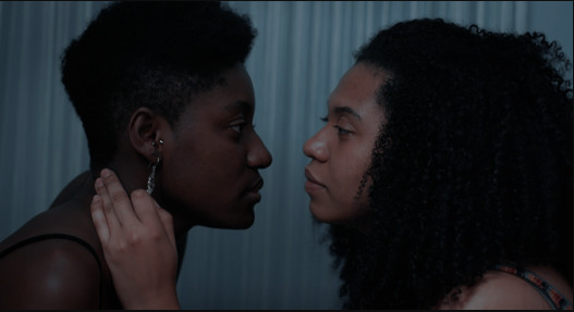 Duas jovens negras, uma de cabelo curto e outra de cabelo na altura dos ombros, estão de perfil, com uma cortina azul acinzentada ao fundo. Elas se olham nos olhos enquanto a jovem de cabelos nos ombros acaricia com uma mão o pescoço da outra