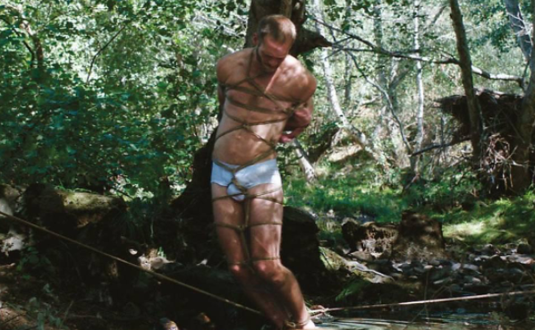Trecho de O ornitólogo. Imagem: Reprodução/O Ornitólogo TA: Em uma floresta, um homem branco, vestindo apenas uma cueca branca, está amarrado a uma árvore.