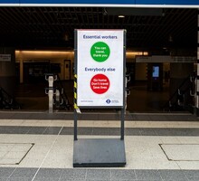 Foto de um cartaz utilizado durante a pandemia. Ele se encontra em uma calçada, na frente de um estabelecimento de transporte. Nele está escrito “trabalhadores essenciais: vocês podem viajar, obrigado” em um círculo verde e “todos os outros: vá para casa. Não viaje. Salve vidas” em um círculo vermelho. O fundo do cartaz é branco e seu suporte é cinza.