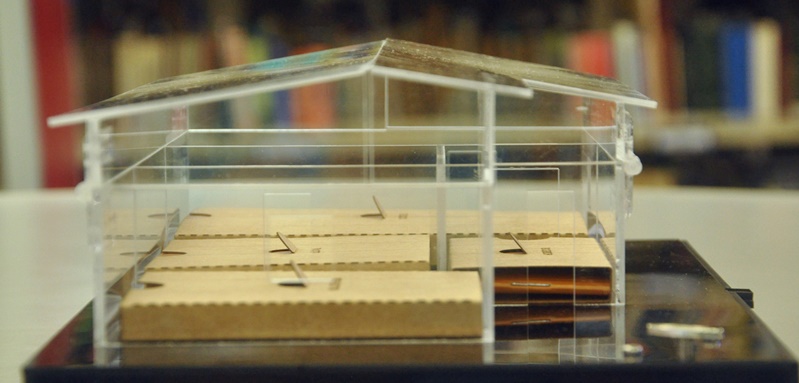 Objeto em forma de casa, em acrílico, com pequenos cadernos dentro de suas divisões. Em segundo plano, desfocada, estante de biblioteca.