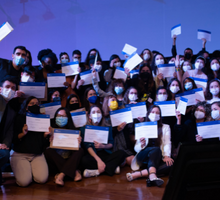 Foto de diversas pessoas segurando seus diplomas. Alguns estão em pé e outros sentados no chão. Todos usam máscara e a maioria olha para a câmera. A luz ao fundo deixa a foto com um aspecto azulado.