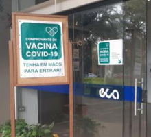 Foto tirada na porta de entrada do prédio principal da ECA. Um mural de madeira está posicionado ao lado de uma porta de vidro. Nele, está pregado um cartaz escrito “Comprovante de vacina covid-19 tenha em mãos para entrar!”. No chão, há uma planta. Pelo reflexo da porta são vistas algumas árvores.