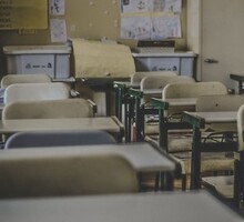 Foto de uma sala de aula vazia. As carteiras são de plástico e aparentam ser gastas. A cor do plástico fica entre o branco e o amarelo, enquanto o metal que o sustenta é verde. Ao fundo, há cartazes feitos pelos estudantes colados na parede. 