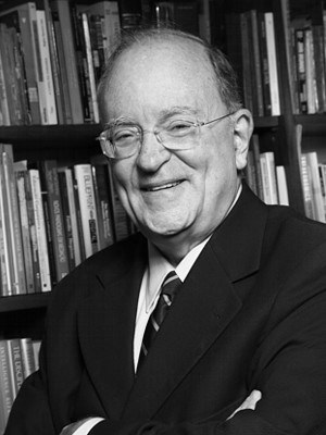 Foto em preto e branco de um homem branco sorrindo. Ele é parcialmente calvo, usa oculos de grau de haste transparente. Na foto, ele veste, paleto, camisa e gravata. Ao fundo, uma estante de livros