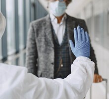 Foto de um agente sanitário barrando um homem aparentemente idoso no corredor de um aeroporto. O agente está com roupas de proteção brancas, uma luva de borracha azul e mostra a palma da mão para o homem, parando-o. O senhor está com um casaco cinza e uma máscara. Ele segura a alça de uma mala.