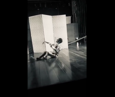 Print de tela mostra imagem preto e branco do ensaio da peça. Um homem está sentado no palco, com as pernas ligeiramente dobradas e o tronco reclinado para trás. Ele estica e ergue um dos braços, enquanto o outro está semi-flexionado. Ao fundo, um biombo, na cor branca.