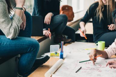 Foto colorida em ambiente interno, de jovens conversando e fazendo projetos em papel