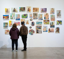 Foto de duas pessoas de costas observando a exposição. A da esquerda é uma mulher, ela veste um casaco roxo e calças pretas. A da direita é um homem, ele usa um casaco marrom e calças pretas. A obra observada é um conjunto de pinturas coloridas. A maioria das telas é retangular, mas quatro são circulares.