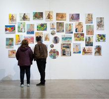  Foto de duas pessoas de costas observando a exposição. A da esquerda é uma mulher, ela veste um casaco roxo e calças pretas. A da direita é um homem, ele usa um casaco marrom e calças pretas. A obra observada é um conjunto de pinturas coloridas. A maioria das telas é retangular, mas quatro são circulares.