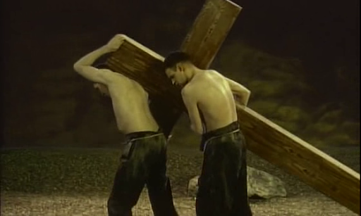 Cena do filme O Jardim. Duas pessoas brancas, sem camisa, com calças pretas, carregando uma enorme cruz de madeira.  