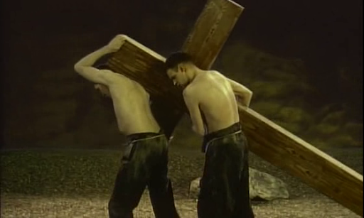 Cena do filme O Jardim. Duas pessoas brancas, sem camisa, com calças pretas, carregando uma enorme cruz de madeira.