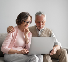 Foto de um casal de idosos. O homem e a mulher estão sentados em um sofá e utilizando juntos um laptop cinza. Ela veste uma blusa rosa e uma calça cinza, enquanto ele usa roupas inteiramente beges. Ele a abraça, e a mão dele está em cima do ombro dela. Os dois sorriem.