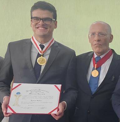 Foto de um jovem de pele branca, cabelos castanhos escuros e óculos de armação retangular posando com um diploma e uma medalha dourada no pescoço. Ele está de pé, usa terno, gravata azul marinho e camisa branca. Ele sorri. Ao lado dele, um homem idoso e careca de terno azul marinho, camisa branca com estampa quadriculada, gravata azul e medalha dourada no pescoço, posa com expressão neutra.