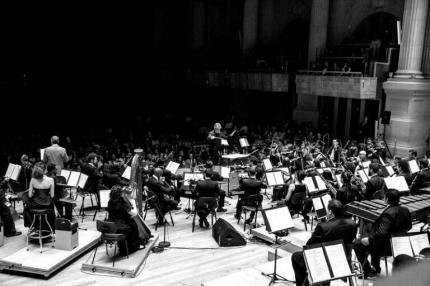 Foto de uma orquestra fotografada do fundo do palco, onde é possível ver diversos instrumentistas, o regente de frente e parte da plateia, ao fundo.