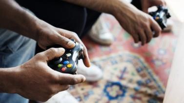 Foto das mãos negras de duas pessoas, cada uma segurando um controle de video game. Ao fundo, desfocado, um tapete no chão.