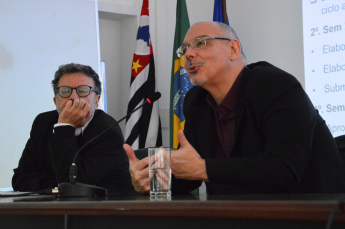 Foto de dois homens sentados em uma mesa. à esquerda, um homem de cabelos levemente grisalhos, usando oculos com haste escura, um terno preto e uma camisa branca, está com o queixo apoiado em uma das mãos. à Direita, um homem calvo, de barba rala, usando oculos, um paleto preto e uma camisa escura fala ao microfone e gesticula com as mãos. Ao fundo, uma parede branca e as bandeiras do estado de são paulo, do brasil e da USP.