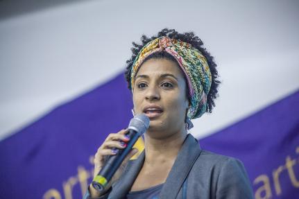 Foto de Marielle Franco, uma mulher negra, falando ao microfone. Ela usa uma faixa colorida nos cabelos crespos e curtos, um terno cinza e suas unhas estão pintadas de roxo.