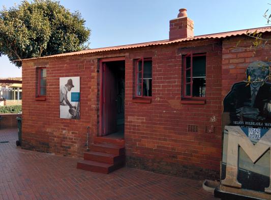Foto da parte de trás da Casa de Mandela, ponto turístico de Joanesburgo. Casa de um andar revestida de tijolinhos vermelhos e com uma chaminé. Do lado esquerdo da estrutura há uma janela vermelha e estreita com três divisões horizontais e um pôster com a fotografia de uma pessoa de chapéu. Do lado direito, duas das mesmas janelas. Ao centro, há um lance de quatro degraus para acessar uma porta vermelha, que está aberta. À frente da casa do lado direito da imagem, há uma estrutura/estátua de Nelson Mandela e ao fundo, à esquerda, aparece sobre a casa a copa de uma árvore.