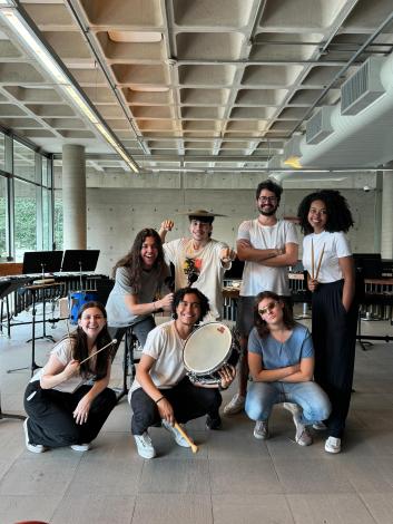 Foto de um grupo de sete pessoas posando, com algumas segurando instrumentos de percussão como baquetas e caixa. São quatro pessoas de pé e três agachadas, a maioria vestindo camisetas brancas. Estão numa sala com diversos instrumentos de percussão. O chão é cinza, a parede da esquerda é de vidro e o teto tem formas quadriculadas em cimento.