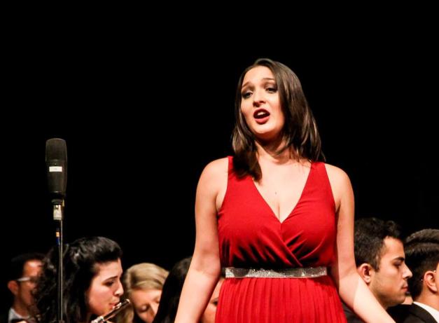 Na foto, uma mulher branca de cabelos castanhos e lisos um pouco abaixo dos ombros está cantando. Ela usa vestido vermelho com decote cruzado, cinto com pedras brilhantes. À esquerda, um microfone. Atrás dela, pessoas sentadas, tocando instrumentos, e um fundo escuro.
